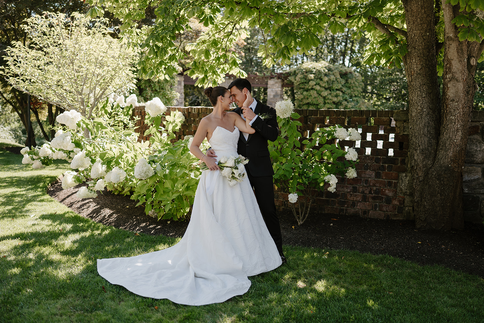 Bride and groom sharing a tender moment by white hydrangeas and a decorative brick wall.